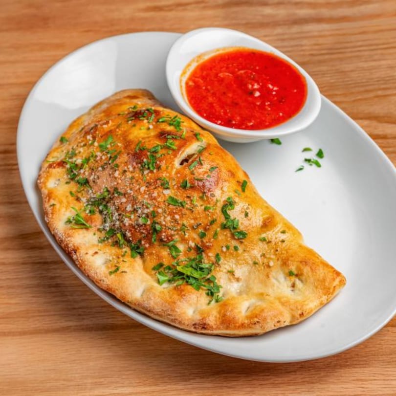 Golden-brown calzone topped with herbs and parmesan, served with a side of red marinara sauce.