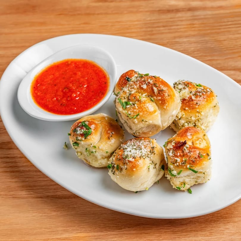 Five golden garlic knots with herbs and parmesan, served with a side of red marinara sauce.