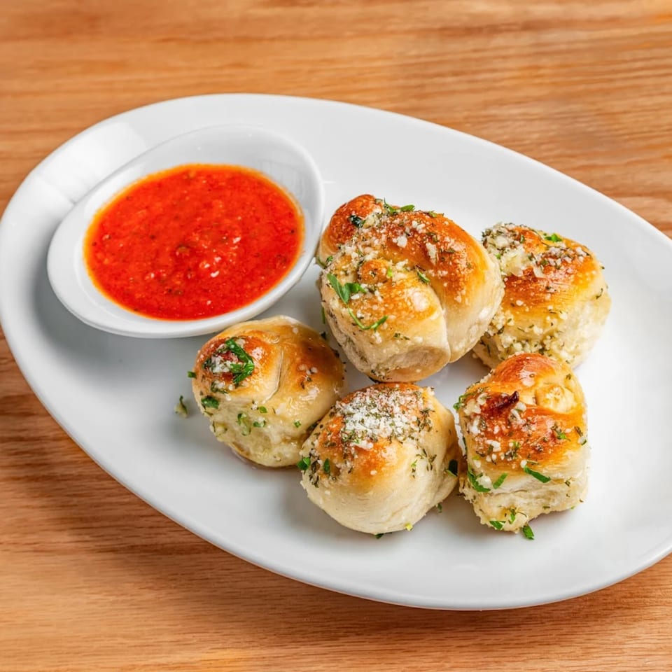 Five golden-brown garlic knots with herbs and Parmesan, served with a side of red marinara sauce.